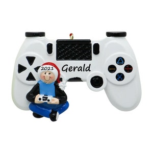 May include: A white video game controller Christmas ornament with a black top and buttons. A figurine of a person wearing a Santa hat and holding a game controller is in front of the controller. The name "Gerald" and the year "2021" are printed on the ornament.