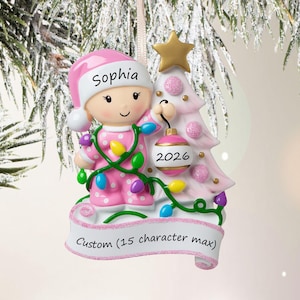 Puede incluir: Un adorno navide&ntilde;o personalizado que muestra a un ni&ntilde;o con pijama rosa de lunares y gorro a juego con el nombre Sophia. El ni&ntilde;o sostiene una bola rosa y blanca con la etiqueta 2026 junto a un &aacute;rbol rosa con estrella dorada y una pancarta.
