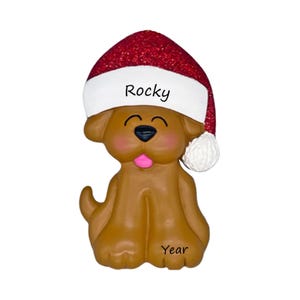 May include: A brown dog ornament wearing a red glittery Santa hat with a white brim. The name Rocky is printed on the brim, and the word Year is printed on the dog's leg. The dog has a black nose, pink tongue, and closed eyes.