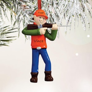 May include: A hanging ornament depicting a figure wearing an orange cap with the word Year, an orange vest with the name Vince, green sleeves, blue pants, and brown boots. The figure holds a brown rifle with a silver barrel, suspended by a red ribbon.