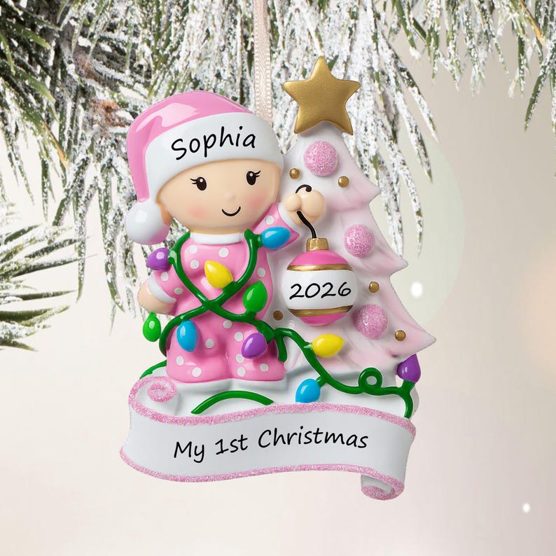 Puede incluir: Un adorno navide&ntilde;o personalizado que muestra a un beb&eacute; con pijama rosa de lunares y gorro a juego con el nombre Sophia. El beb&eacute; sostiene una cadena de luces de colores y una bola rosa marcada con 2026 junto a un &aacute;rbol rosa con estrella.