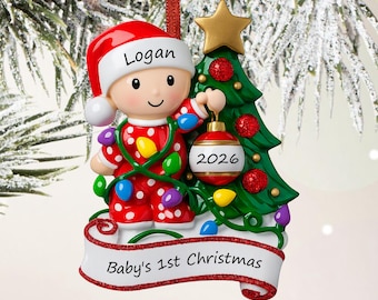 May include: A Christmas tree ornament featuring a baby in red polka-dot pyjamas and a Santa hat with the name Logan, standing next to a green tree with a gold star, a red ornament labelled 2026, and a white banner reading Baby's 1st Christmas.