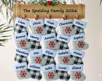Puede incluir: Un adorno para árbol de Navidad con un letrero de madera que dice The Spalding Family 2026 y veinte calcetines en miniatura colgando debajo. Cada calcetín tiene un puño blanco con un nombre, un patrón de cuadros negro y blanco y un copo de nieve rojo.