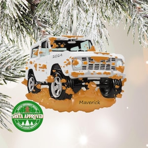 May include: White Ford Bronco Christmas ornament with brown mud splatters, the year 2024 printed on the side, and the name Maverick printed below. The ornament has a green circle with the words "Santa Approved" and "Making Lasting Memories".