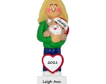 May include: A personalized Christmas ornament featuring a girl with blonde hair holding a cat wearing a Santa hat. The ornament has a red heart with the year 2021 and a white base with the name Leigh Ann.
