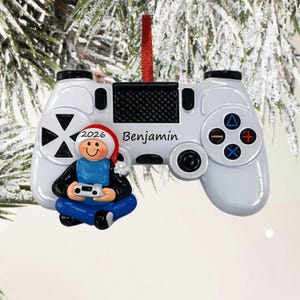 May include: A white video game controller ornament featuring a small figure wearing a Santa hat and blue clothing, holding a miniature controller. The name Benjamin is printed on the controller, and the year 2026 is on the Santa hat brim.