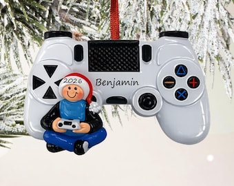 Puede incluir: Un adorno de mando de videojuego blanco con una pequeña figura que lleva un gorro de Papá Noel y ropa azul, sosteniendo un mando en miniatura. El nombre Benjamin está impreso y el año 2026 aparece en el borde del gorro navideño.