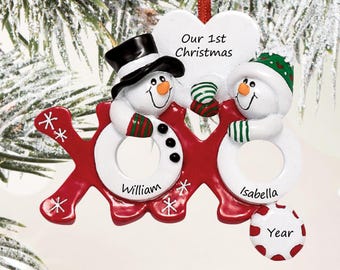 May include: A Christmas ornament featuring two snowmen on a red XOXO base. The left snowman wears a black top hat, the right wears a green hat. A white heart above reads Our 1st Christmas. Names William and Isabella are printed on the snowmen.