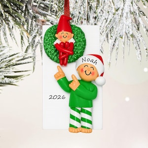 May include: A rectangular white Christmas ornament featuring a green wreath with a red-clad elf and a figure wearing a green shirt, green and white striped pants, and a red hat with the name Noah written on it, with the year 2026 printed below.
