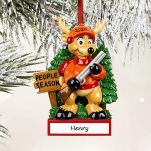 May include: A hanging Christmas ornament featuring a tan deer wearing an orange cap with 2026 printed on it, an orange jacket, and black gloves, holding a rifle. A wooden sign reads PEOPLE SEASON, and a red base displays the name Henry.
