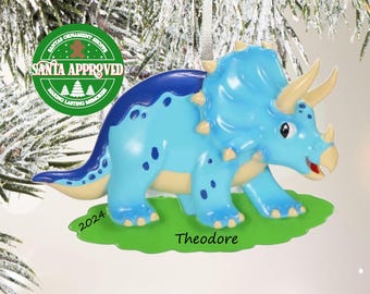 May include: A blue triceratops dinosaur ornament with a dark blue back, cream-colored horns, and a green base. The base features the text 2024 and the name Theodore in black script. A silver ribbon loop is attached to the top for hanging.