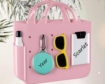 May include: A pink tote bag ornament featuring a side pocket with a white bottle, a teal circular tag with the text Year, yellow sunglasses, and a white rectangular tag with the text Scarlet. The ornament has two handles and a hanging loop.
