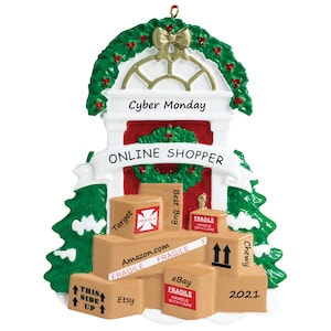 May include: A Christmas ornament shaped like a house with a green wreath and a gold bow. The text "Cyber Monday" and "ONLINE SHOPPER" are displayed. Brown boxes with the logos of Amazon, Etsy, Target, Best Buy, Chewy, and eBay are stacked below. The year 2021 is also displayed.