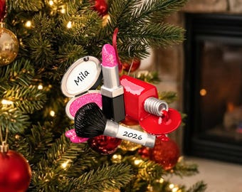 May include: A makeup-themed Christmas ornament featuring a pink glittery lipstick, a black makeup brush with the year 2026 printed on the handle, a red nail polish bottle, and a white compact case with the name Mila printed on the inside lid.