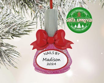 May include: A pink glittery nail polish bottle ornament featuring a silver cap, a large red bow, and a white oval label with the black text NAILS BY Madison 2024. The ornament hangs from a red ribbon against a snowy pine branch background.