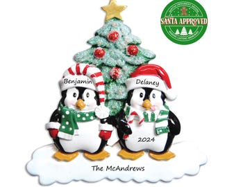 May include: Personalized Christmas ornament featuring two penguins wearing Santa hats and scarves, standing on a snowy base with a Christmas tree in the background. The ornament is inscribed with the names "Benjamin" and "Delaney", the year "2024", and "The McAndrews".