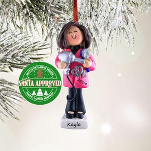 May include: A pink and white Christmas ornament depicting a woman with brown hair holding a hairdryer, scissors, and a comb. The ornament has the text "2024" and "Kayla" on it. The ornament is surrounded by a green circle with the text "Santa Approved" and a gingerbread man.