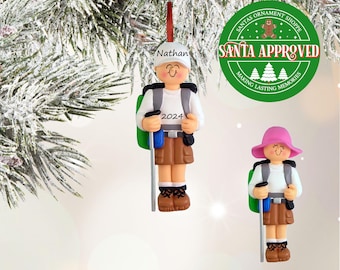 May include: Two personalized Christmas ornaments of hikers with backpacks. The ornament on the left is a male hiker with the name "Nathan" and the year "2024" on his backpack. The ornament on the right is a female hiker with a pink hat. Both ornaments have the text "Santa Approved" on a green circle.
