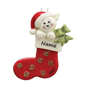 May include: White cat wearing a Santa hat peeking out of a red stocking with gold swirls. The stocking has a white tag with the word "Name" and the year on the bottom.