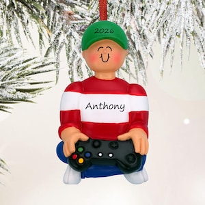 May include: A Christmas tree ornament shaped like a figure wearing a green cap with 2026 printed on it, a red and white striped shirt with the name Anthony printed on the chest, and blue pants, while holding a black video game controller.