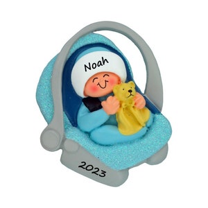 May include: A decorative ornament featuring a baby in a light blue glittery car seat. The baby wears a white hat with the name Noah printed in black. The baby holds a small yellow teddy bear. The base of the car seat is gray with 2023 printed in black.