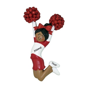 May include: A red and white cheerleader ornament with black hair, a red bow, and pom-poms. The ornament has the text "Name" and "Year" on it.