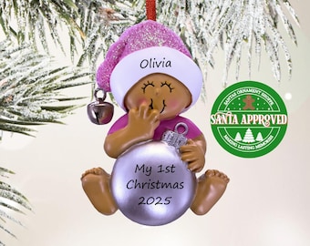 May include: A hanging Christmas ornament shaped like a baby wearing a pink glittery hat with a silver bell. The baby holds a silver ball ornament printed with the text My 1st Christmas 2025. The hat is personalised with the name Olivia in black text.