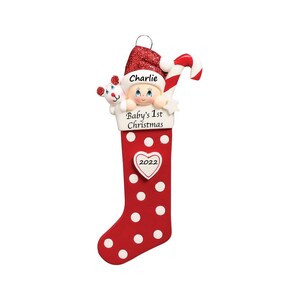 May include: Red and white polka dot stocking ornament with a baby peeking out of the top. The stocking has a white heart with the year 2022 on it. The baby is wearing a Santa hat and holding a stuffed animal. The text "Charlie" and "Baby's 1st Christmas" is on the stocking.