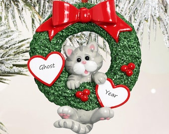 May include: A Christmas tree ornament featuring a grey tabby cat peeking through a green glittery wreath. The wreath is topped with a red bow and decorated with red berries and two white heart-shaped plaques labeled Ghost and Year.