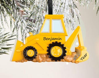 May include: A yellow construction vehicle ornament with black wheels and a brown base. The side of the vehicle features the name Benjamin in black script, and the rear bucket area has the word Year printed in black. It hangs from a black ribbon.
