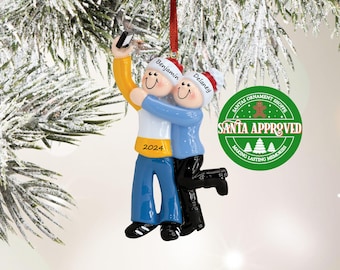 Puede incluir: Un adorno navideño con dos figuras abrazándose y haciéndose un selfie. Las figuras llevan gorros de Papá Noel y ropa azul y amarilla. El adorno tiene los nombres "Benjamin" y "Delaney" en los gorros y el año "2024".