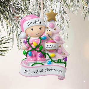 Puede incluir: Un adorno navide&ntilde;o personalizado que muestra a un beb&eacute; con pijama rosa de lunares y un gorro a juego con el nombre Sophia. El beb&eacute; sostiene una bola rosa con la etiqueta 2026 junto a un &aacute;rbol rosa con una estrella dorada. Una cinta dice Baby's 2nd Christmas.