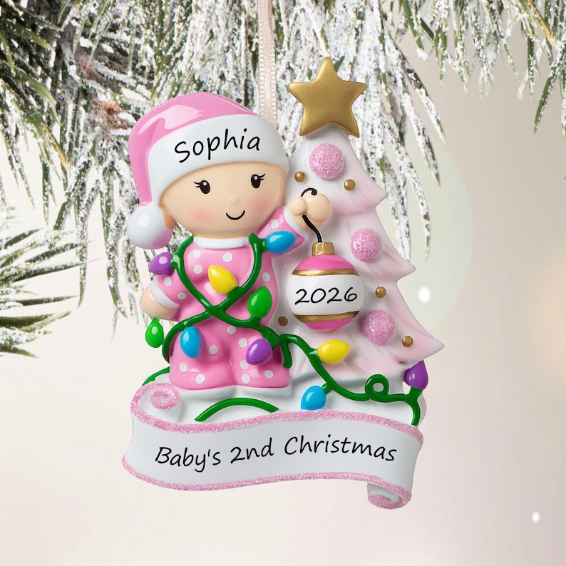 Puede incluir: Un adorno navide&ntilde;o personalizado que muestra a un beb&eacute; con pijama rosa de lunares y un gorro a juego con el nombre Sophia. El beb&eacute; sostiene una bola rosa con la etiqueta 2026 junto a un &aacute;rbol rosa con una estrella dorada. Una cinta dice Baby's 2nd Christmas.