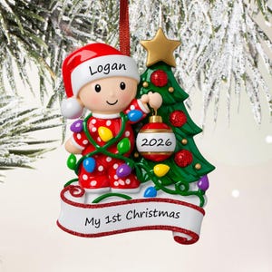 Puede incluir: Un adorno de &aacute;rbol de Navidad con un ni&ntilde;o en pijama rojo de lunares y gorro de Pap&aacute; Noel con el nombre Logan. El ni&ntilde;o sostiene un adorno rojo con la etiqueta 2026 junto a un &aacute;rbol verde con una estrella dorada. Una cinta blanca dice My 1st Christmas.