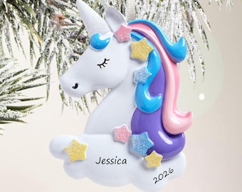 Puede incluir: Un adorno de unicornio blanco con un cuerno brillante, con una melena multicolor en azul, morado y rosa. El unicornio descansa sobre una base de nube blanca con el nombre Jessica y el año 2026 impresos en negro, decorado con pequeñas estrellas.