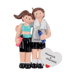May include: A decorative ornament featuring two figures in athletic wear, one holding a pink water bottle and the other a blue water bottle with a white towel around their neck. The year 2022 is printed on the shirt, and a heart displays the names Maverick & Charlotte.