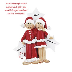 May include: A Christmas ornament featuring two figures wearing red and white Santa suits with matching hats. The figures stand before a large white star labelled Year, with two smaller stars on the sides labelled Name 1 and Name 2 respectively.
