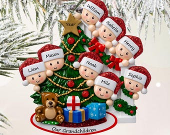 Puede incluir: Un adorno de árbol de Navidad con siete figuras de dibujos animados con gorros rojos brillantes con los nombres Liam, Mason, William, Isabella, Henry, Noah, Mila y Sophia. Un oso de peluche y dos regalos tienen el texto Our Grandchildren.