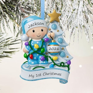 Puede incluir: Un adorno navide&ntilde;o con un beb&eacute; en pijama azul y gorro a juego con el nombre Jackson, junto a un &aacute;rbol blanco con detalles de purpurina azul y una estrella dorada. El beb&eacute; sostiene una bola azul con el a&ntilde;o 2026 y una guirlande de luces.