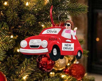 May include: A red car-shaped Christmas ornament featuring a figure wearing sunglasses in the driver seat. The car is labeled with the name Olivia, the year 2026, and a sign on the door that reads NEW DRIVER in black text on a white background.