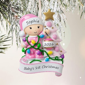 Puede incluir: Un adorno navide&ntilde;o con un beb&eacute; en pijama rosa de lunares y gorro a juego con el nombre Sophia, sosteniendo una bola rosa con la etiqueta 2026 junto a un &aacute;rbol rosa con una estrella dorada. Una pancarta en la base dice Baby's 1st Christmas.