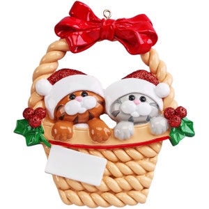 May include: A hanging Christmas ornament featuring a tan woven basket with a large red bow at the top. Inside the basket are two cats, one orange tabby and one gray tabby, both wearing red glittery Santa hats. Green holly leaves with red berries decorate the sides, and a blank white rectangular plaque is attached to the front for personalization.