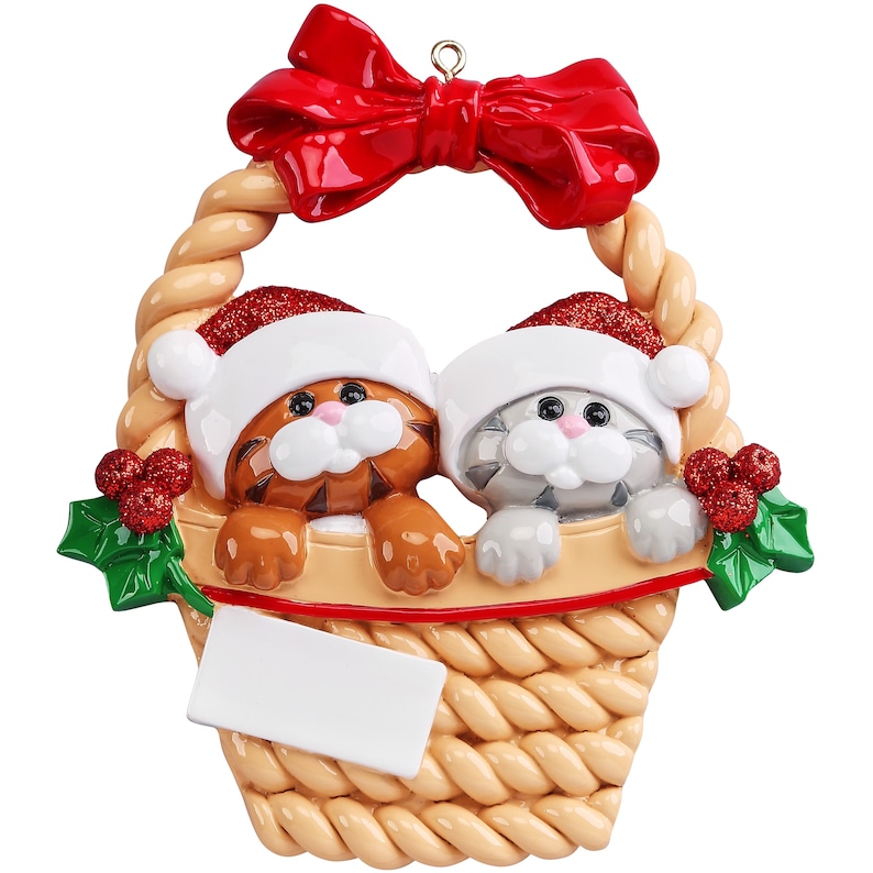 May include: A hanging Christmas ornament featuring a tan woven basket with a large red bow at the top. Inside the basket are two cats, one orange tabby and one gray tabby, both wearing red glittery Santa hats. Green holly leaves with red berries decorate the sides, and a blank white rectangular plaque is attached to the front for personalization.