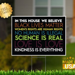 May include: Black yard sign with the text "In this house we believe Black Lives Matter, Women's Rights are Human Rights, No Human is Illegal, Science is Real, Love is Love, Kindness is Everything". The sign is on a green lawn with a gold "Made in USA" sticker in the bottom right corner.