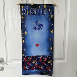 May include: A vertical fabric wall organiser featuring a blue gradient background with yellow stars. The word DISNEY is printed at the top in blue. It includes a central wizard hat icon, a red Mickey Mouse head, and a bottom pocket with a repeating pattern.
