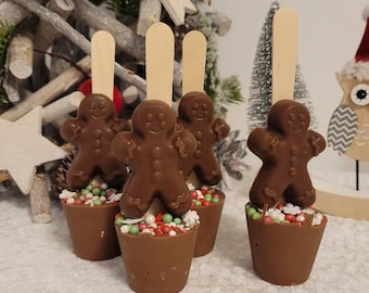May include: Four chocolate gingerbread man hot chocolate stirrers with wooden sticks. Each stirrer is in a small brown cup filled with colorful sprinkles. The background includes a Christmas tree and decorative elements.