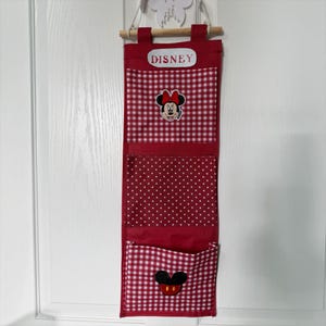May include: A red fabric hanging wall organiser with three pockets, suspended from a wooden dowel. The top pocket features a red and white gingham pattern with a Minnie Mouse patch and a white label reading DISNEY. The middle pocket is red with white polka dots, and the bottom pocket is red and white gingham with a Mickey Mouse icon patch.