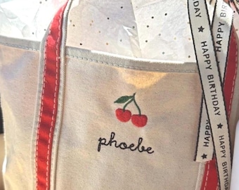 Könnte beinhalten: Eine cremefarbene Canvas-Tragetasche mit roten vertikalen Streifen, einem gestickten Kirschmotiv und dem Namen "phoebe" in schwarzer Schreibschrift. Ein cremefarbenes Band mit dem Aufdruck "HAPPY BIRTHDAY" ist am Griff befestigt.