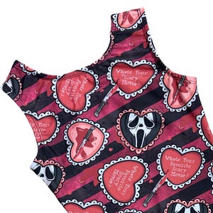 May include: A sleeveless garment featuring a repeating pattern of pink hearts with white lace-style borders on a black and dark red striped background. The hearts contain the text "What's Your Favorite Scary Movie?" alongside illustrations of knives and ghost-like masks.