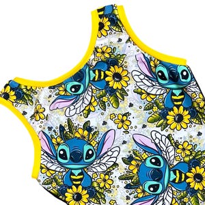 Op de afbeelding: Een mouwloze top met een herhaald patroon van een blauw stripfiguur met grote ogen, bijachtige vleugels en gele en groene bloemaccenten. De top heeft een witte achtergrond en gele rand.
